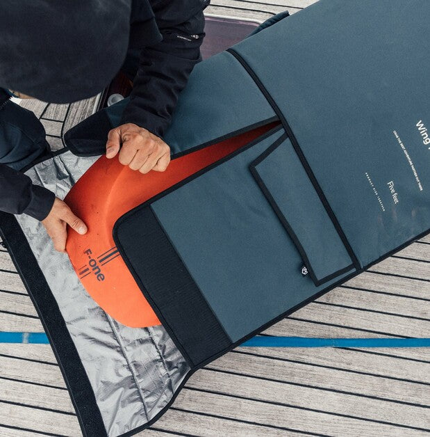 Person holding an orange and black surfboard against a blue mat on a wooden deck.