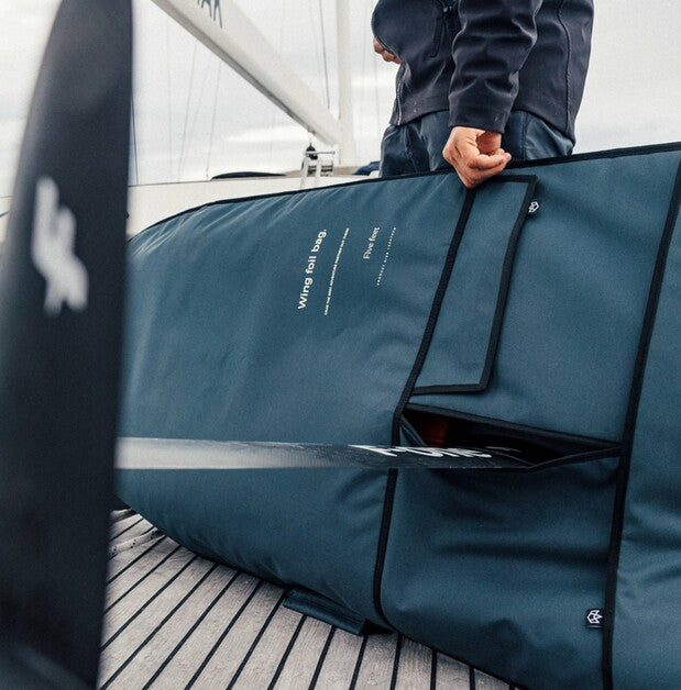 A large dark teal Wind Sail Bag with black trim and white printed text, being held by a person on a boat deck next to a black sail sleeve.