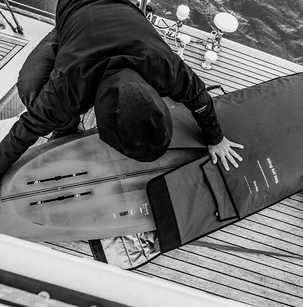 A person in a black hoodie and cap is lying on a wooden boat deck next to a surfboard and a black carrying bag. The photo is in black and white.
