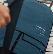 A dark teal wing foil bag with black trim and white printed text, shown outdoors on a wooden deck with a hand holding a strap in the foreground.
