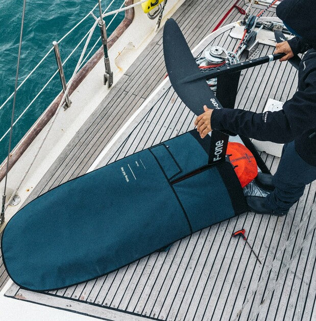 A person on a sailboat deck packing an F-One foil board into a dark teal travel bag.