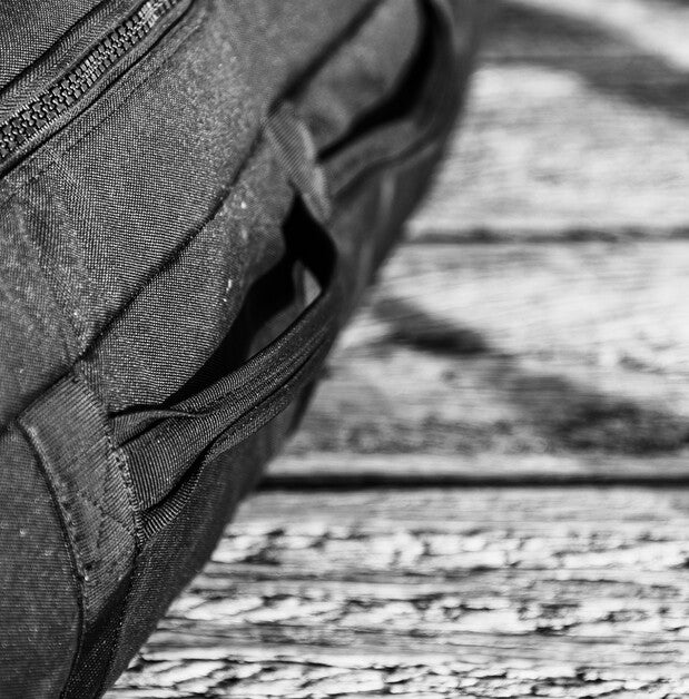 Close-up of a dark gray fabric bag with a zipper and side pocket, resting on a weathered wooden surface in black and white.