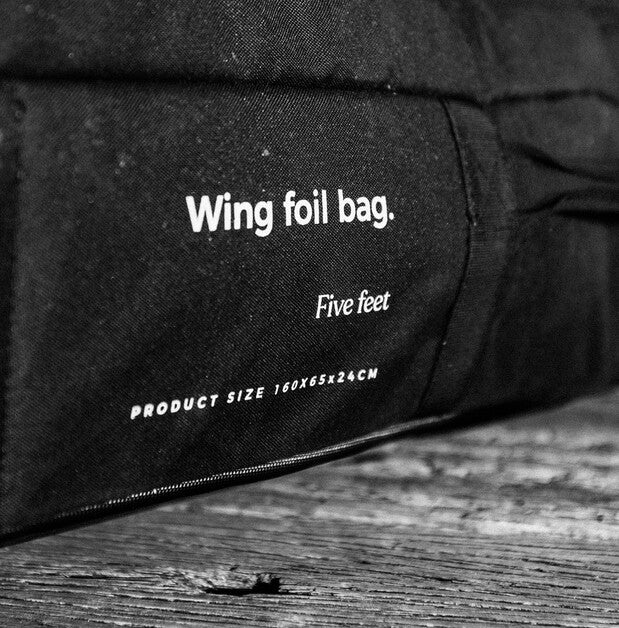 A close-up black fabric pouch with white printed text reading 'Wing foil bag.', 'Five feet', and 'PRODUCT SIZE 160x65x24CM', resting on a rustic wooden surface.