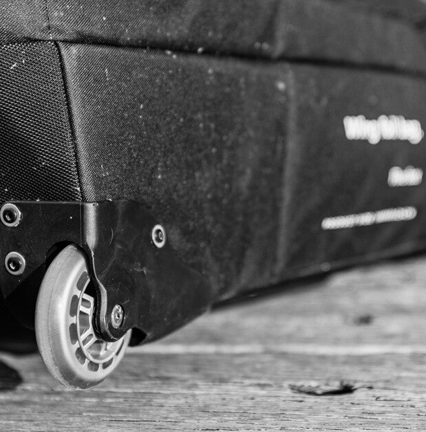 Close-up of a black wheeled suitcase or duffel bag corner with a visible wheel and metal hardware, photographed on a rustic wooden surface.