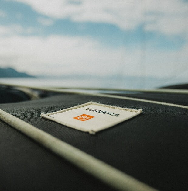 A close-up shot of a black textured fabric surface with a white stitched border and a sewn-on brand label reading MANERA, with a blurred outdoor background of water and sky.