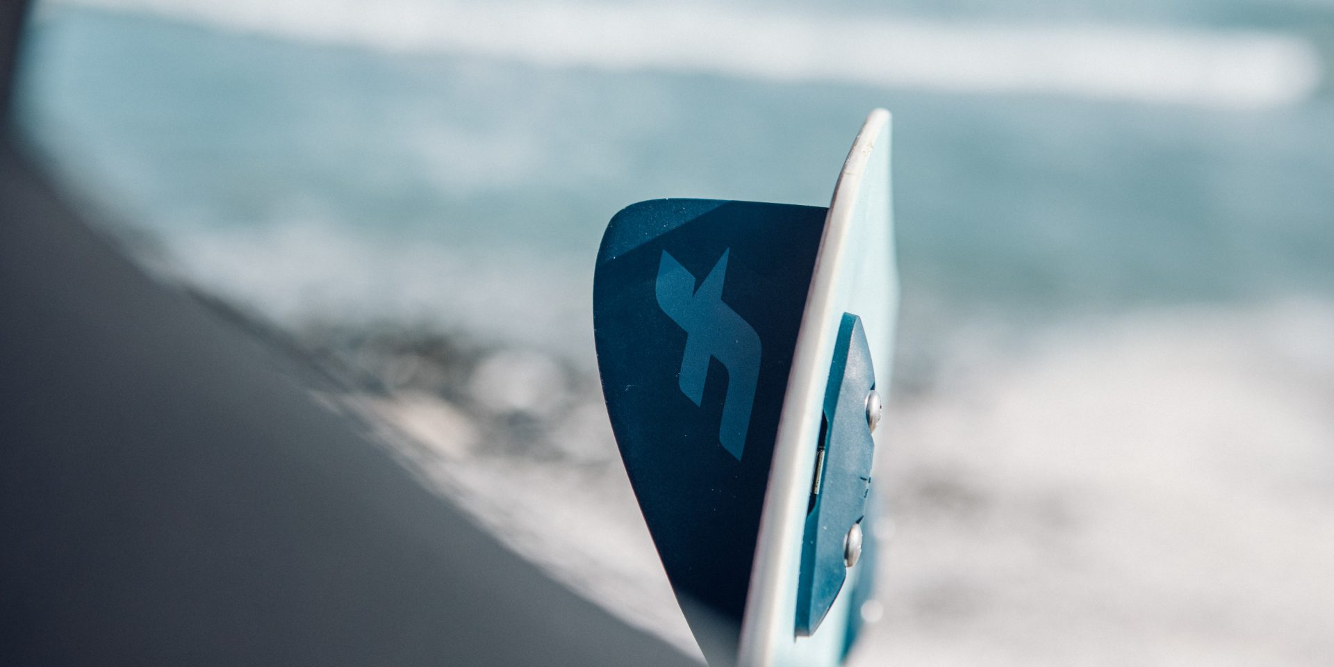 Close-up of a dark blue surf fin with an S-shaped logo, mounted on the underside of a light blue board, with the beach and ocean in the background.