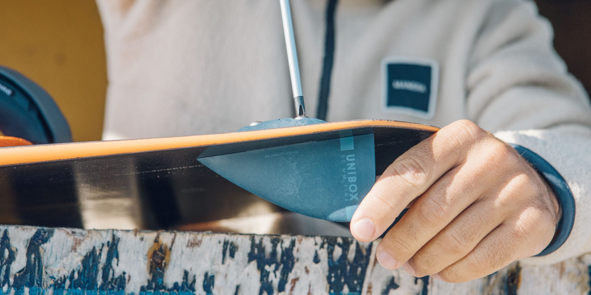 A close-up of a person's hand holding a dark teal snowboard, showing the 'UNIQEX' brand logo on the base, while wearing a light beige fleece jacket.